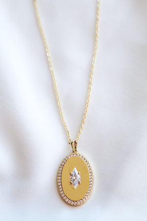 The Fique Necklace by Kinsey Designs is a gold-plated 32-inch chain with an oval cubic zirconia pendant bordered by small diamonds, elegantly displayed on white fabric.