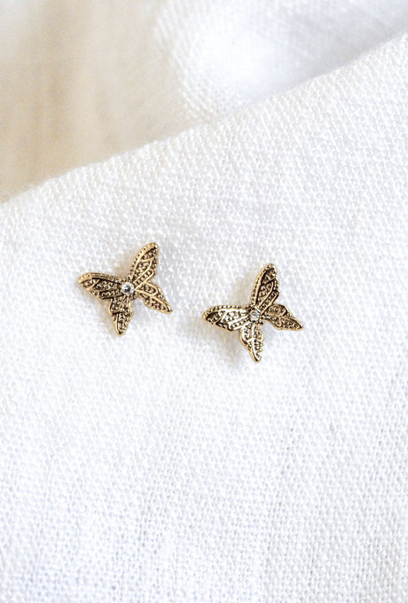 The Fleur gold plated butterfly-shaped earrings by Kinsey Designs Archives are elegantly displayed on a textured white fabric background, showcasing their sophisticated jewelry design.