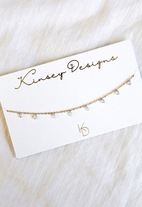 The Clare Choker by Kinsey Designs Archives features a delicate gold-plated 14" chain with small dangling cubic zirconia crystals, presented on a branded card for an elegant, dainty look.