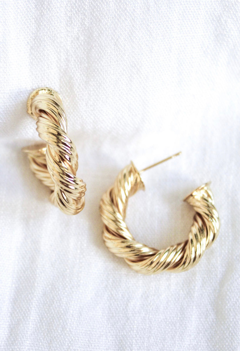 Kinsey Designs' Twisted Micayla Hoop earrings are elegantly displayed on a white textured fabric background, highlighting the timeless appeal of these twisted gold hoops.
