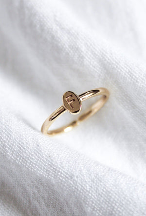 The Kinsey Initial Ring by Kinsey Designs features a small oval gold face engraved with the letter "F," shown on a white fabric background—a stunning personalized jewelry piece.