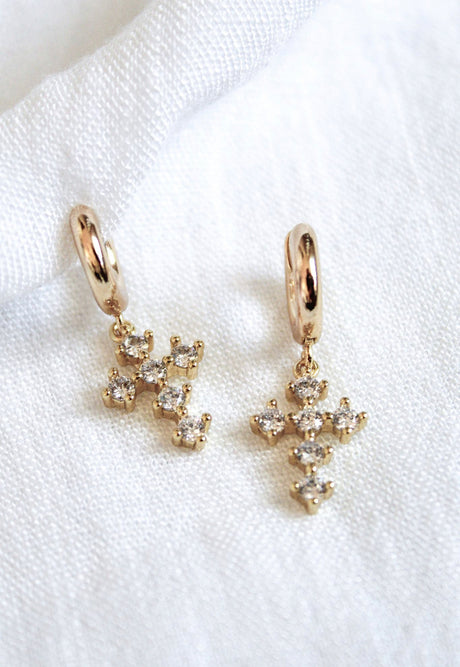 The Tori Huggie - Clear by Kinsey Designs Archives features 1.25-inch gold-filled hoops with dangling cross-shaped pendants adorned with sparkling cubic zirconia crystals, shown on white fabric.