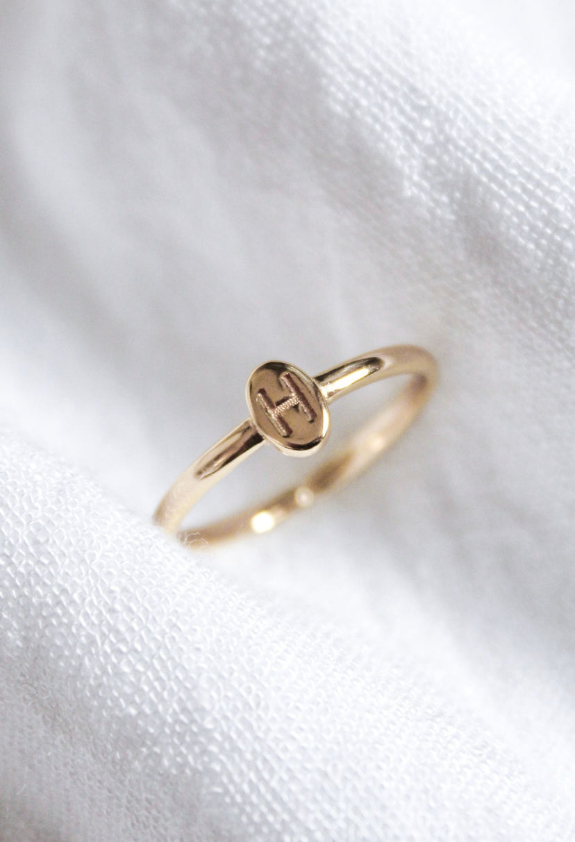 The Kinsey Initial Ring by Kinsey Designs, engraved with an “H,” is shown on textured white fabric—an elegant, personalized jewelry piece ideal for daily wear.