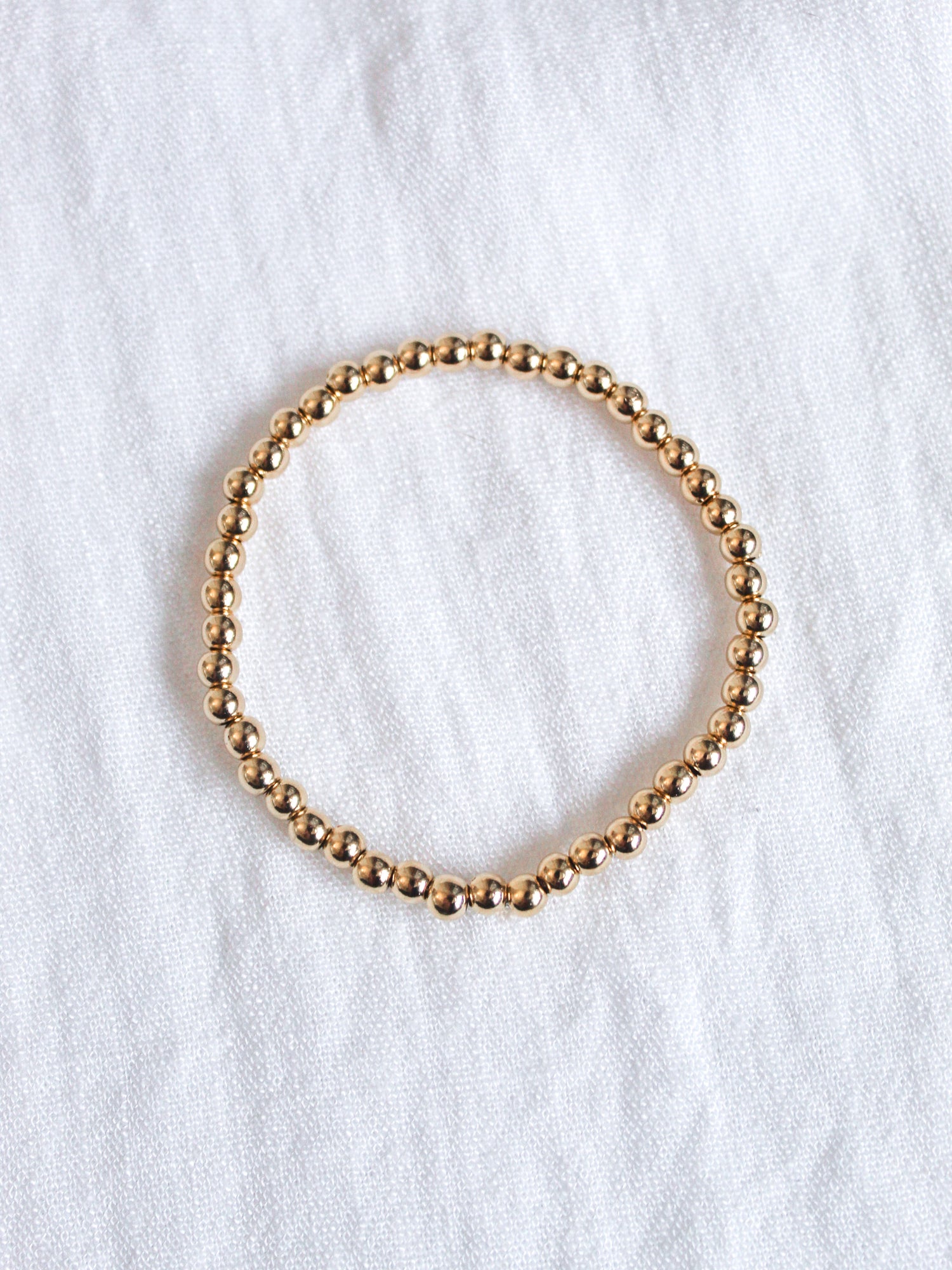 The Kinsey Designs Liam Stacking Bracelet, available in multiple sizes, features gold-filled beads and an adjustable stretch fit, shown arranged in a circle on a white fabric background.