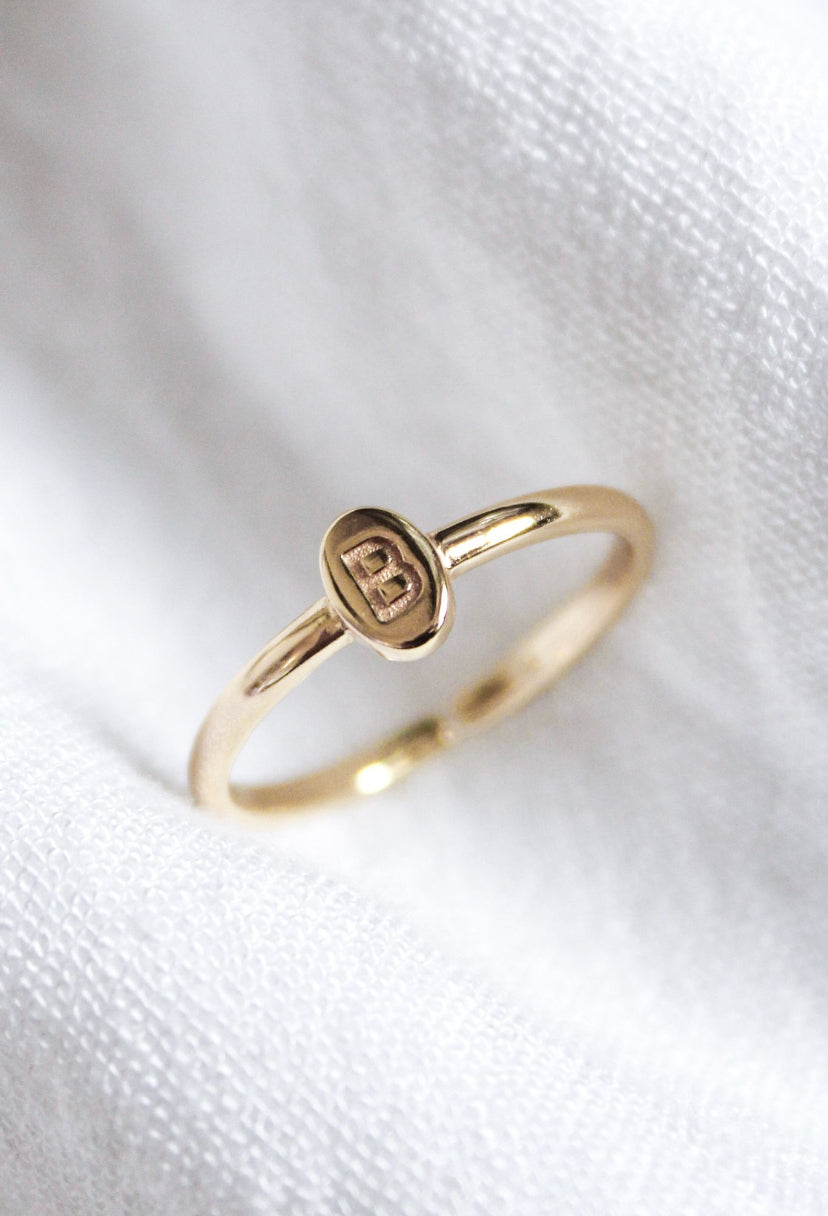 The Kinsey Initial Ring by Kinsey Designs features an oval gold plate engraved with the letter "B," displayed on white fabric—a timeless, personalized jewelry piece.
