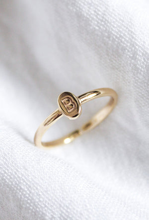 The Kinsey Initial Ring by Kinsey Designs features an oval gold plate engraved with the letter "B," displayed on white fabric—a timeless, personalized jewelry piece.