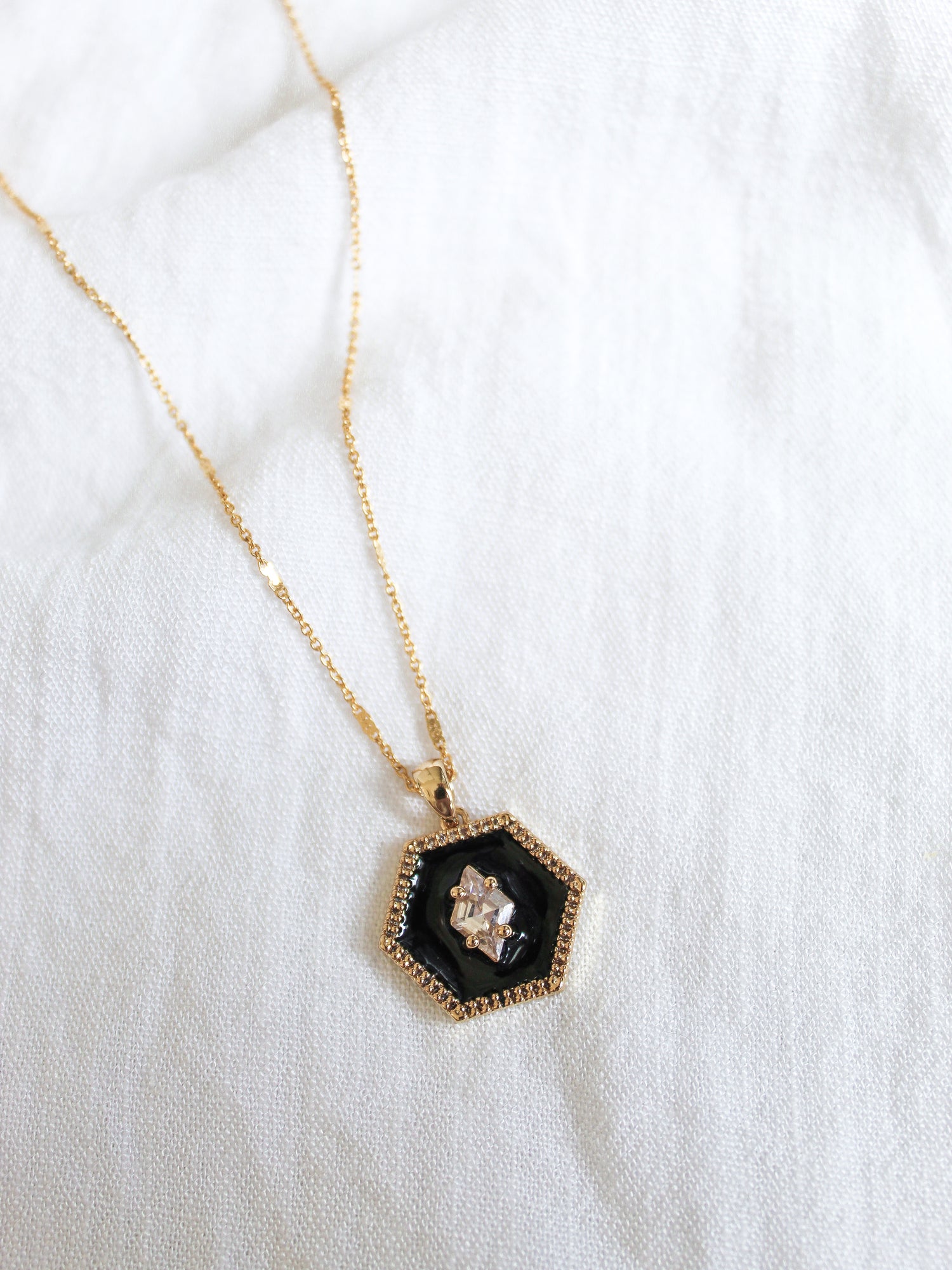 The Nova Necklace by Kinsey Designs Archives features a hexagonal black gold plated pendant with a clear cubic zirconia gemstone, displayed elegantly on white fabric.