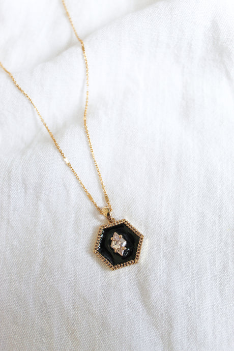 The Nova Necklace by Kinsey Designs Archives features a hexagonal black gold plated pendant with a clear cubic zirconia gemstone, displayed elegantly on white fabric.