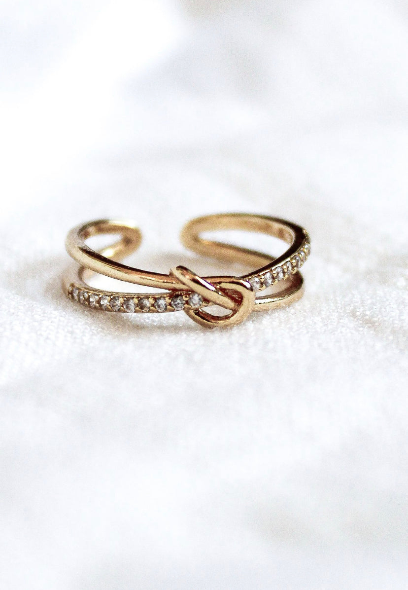 The Kinsey Designs Archives Knotty Ring is a gold-plated, adjustable ring with a knot design and small gemstones, fitting sizes 7-9. Shown on soft white fabric.