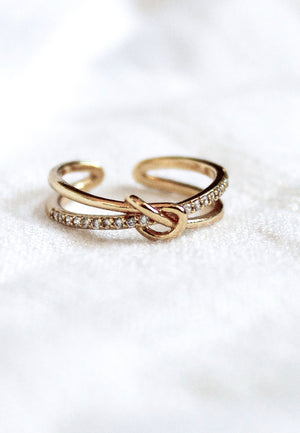 The Kinsey Designs Archives Knotty Ring is a gold-plated, adjustable ring with a knot design and small gemstones, fitting sizes 7-9. Shown on soft white fabric.