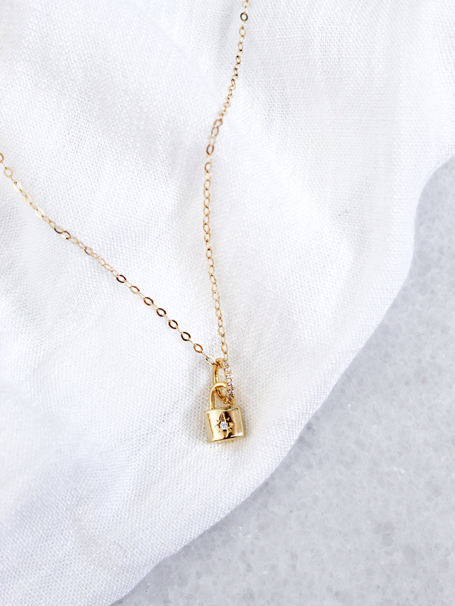 The Blaire Lock Necklace by Kinsey Designs Archives showcases a delicate 16k gold filled chain and a gold plated brass padlock charm, displayed elegantly on a white fabric background.