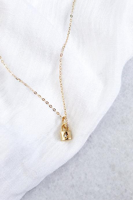 The Blaire Lock Necklace by Kinsey Designs Archives showcases a delicate 16k gold filled chain and a gold plated brass padlock charm, displayed elegantly on a white fabric background.
