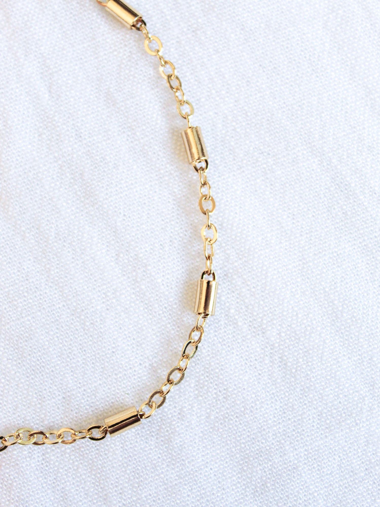 The gold chain bracelet with cylindrical beads rests on a white textured fabric, paired with the Kinsey Designs Archives Azure Anklet - Gold Filled for added elegance.