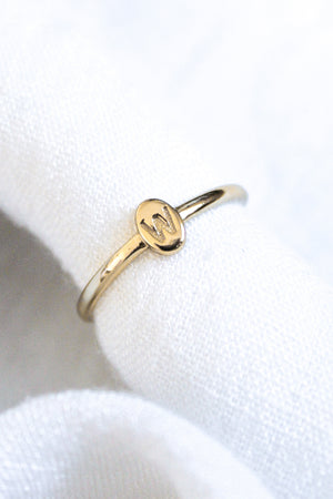 The Kinsey Initial Ring by Kinsey Designs features a gold round bezel engraved with the letter "W," displayed on white textured fabric—an elegant, personalized jewelry piece.