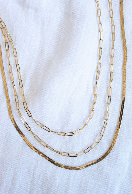 The Wren Chain Layer by Kinsey Designs Archives, a three-layered 16k gold-plated brass necklace with rectangular links, is elegantly displayed on a white fabric background.