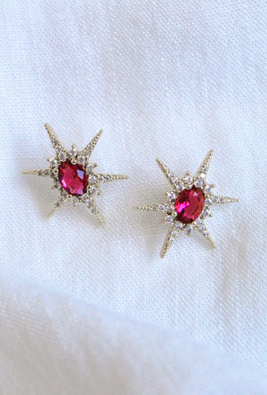 The Faye Stud earrings by Kinsey Designs feature a gold-filled starburst design with red gemstones and sparkling cubic zirconia, shown on white fabric.
