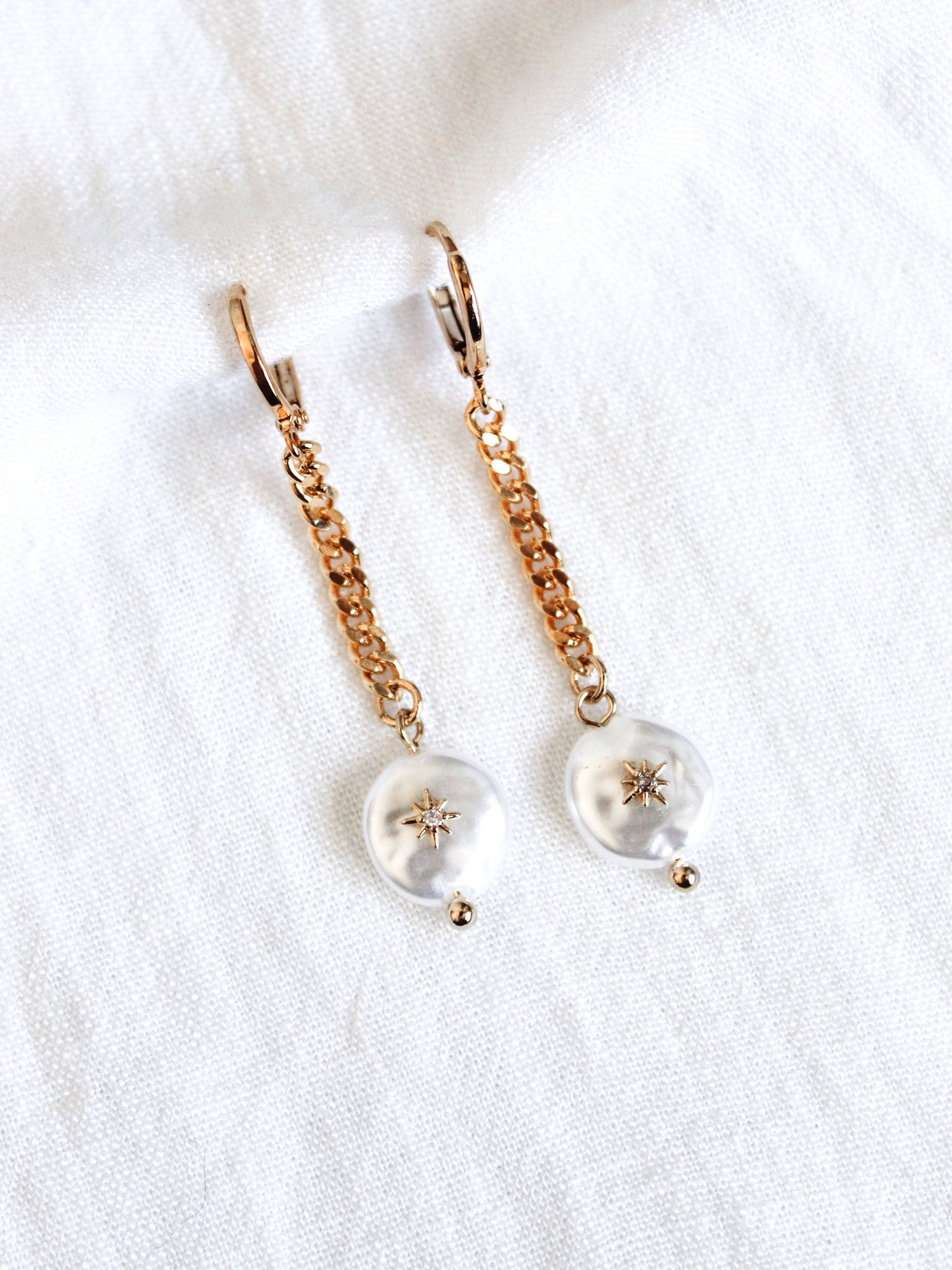 The Harper Drop earrings by Kinsey Designs Archives feature gold plated brass chains, 1.5 inches long, with pearl-like dangles and star accents, displayed on a white fabric background.