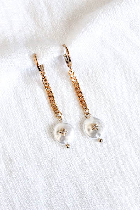 The Harper Drop earrings by Kinsey Designs Archives feature gold plated brass chains, 1.5 inches long, with pearl-like dangles and star accents, displayed on a white fabric background.