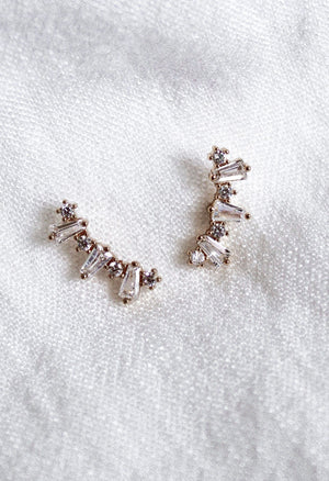 The Irace Post earrings by Kinsey Designs Archives feature small cubic zirconia stones in a curved line, displayed on white textured fabric.