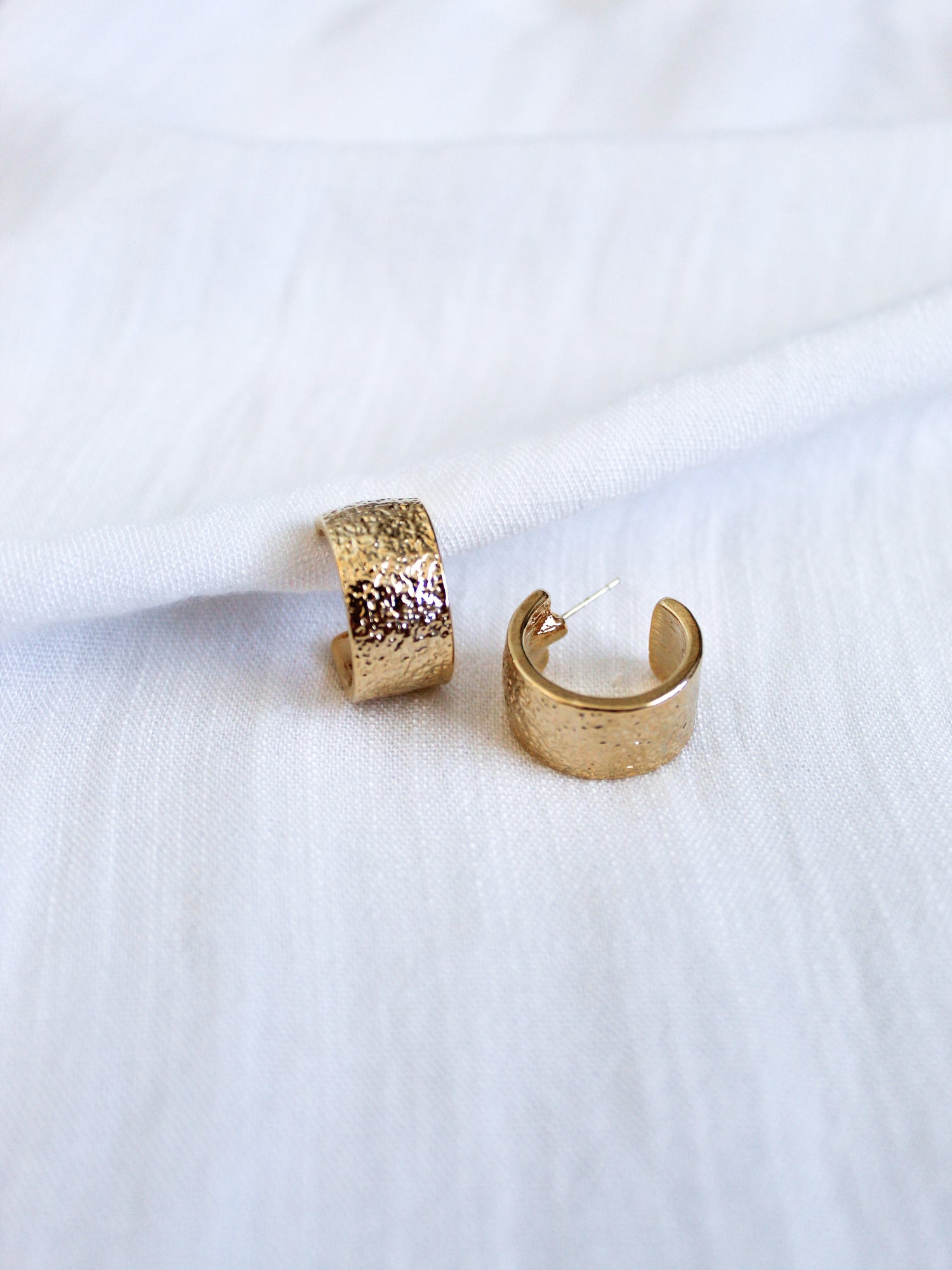 Two Fawn Hoop gold-plated brass earrings by Kinsey Designs Archives, featuring a textured finish, rest elegantly on a white fabric background.