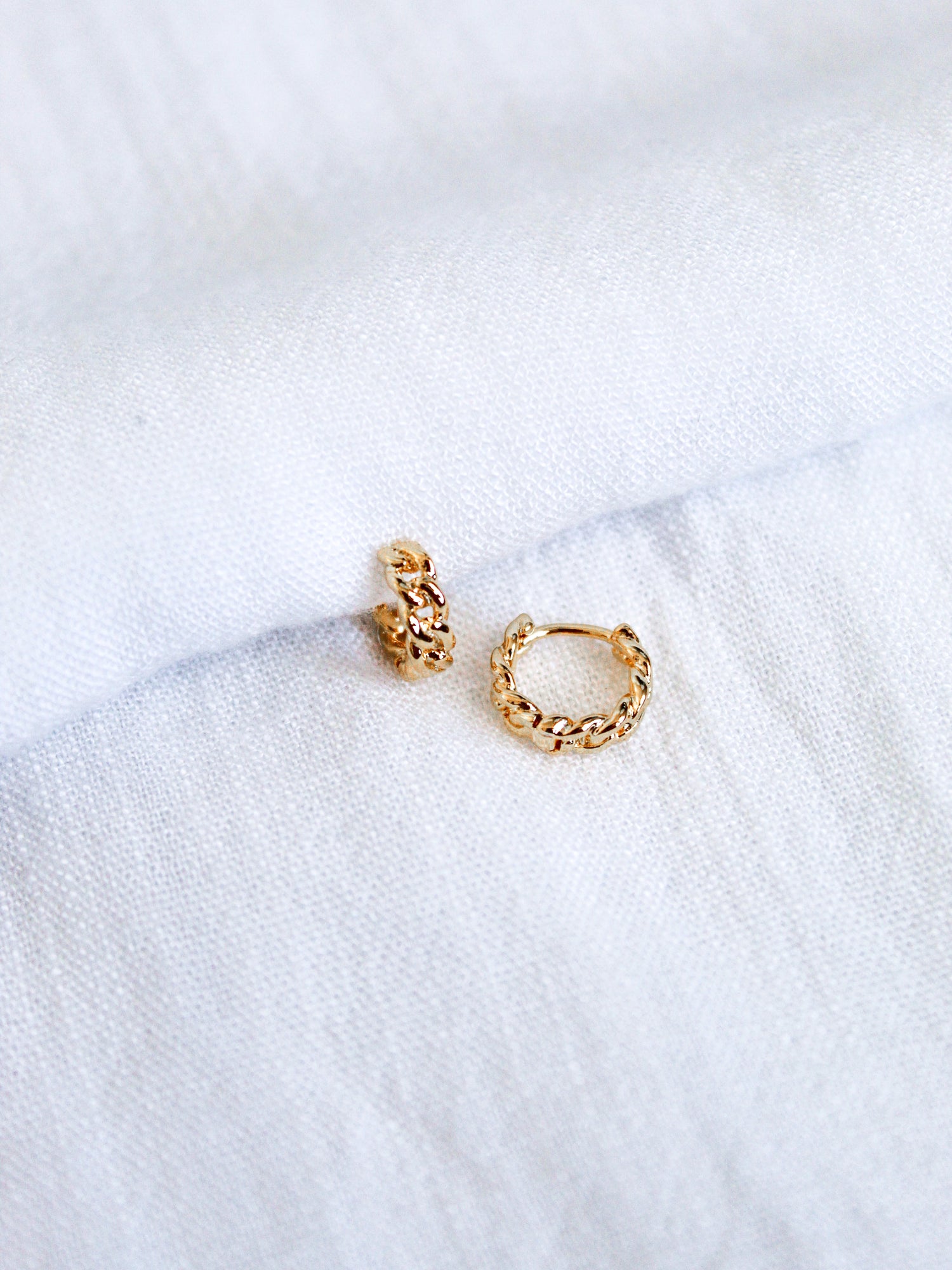 The Ivy Huggie by Kinsey Designs Archives features two small 16k gold plated brass hoops with a braided design, shown on a white fabric background.