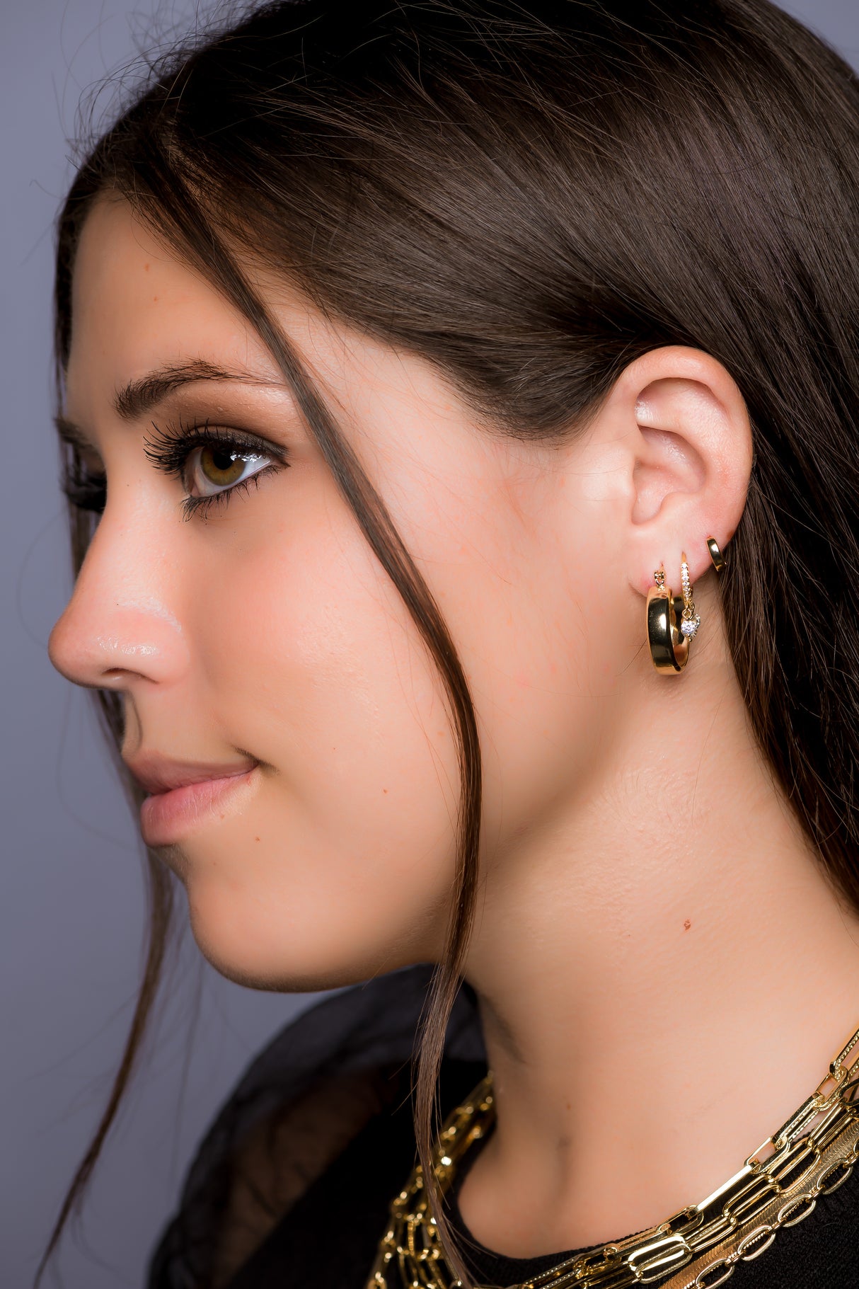 A woman in profile with long brown hair wears Kinsey Designs Archives’ Doreen gold filled necklace and gold hoop earrings, looking left.