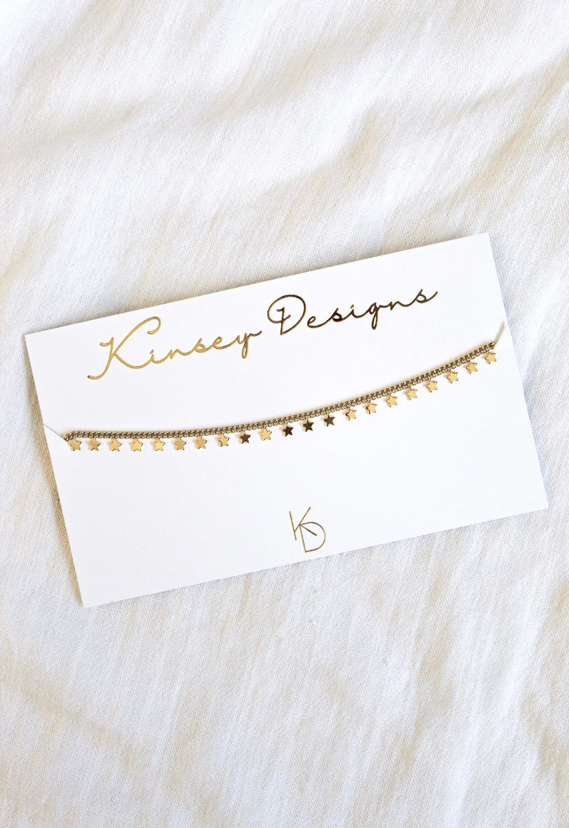 The Starry Choker by Kinsey Designs Archives, a gold-plated 14” necklace with a star charm, is displayed on a white branded card against white fabric for an elegant, stylish look.