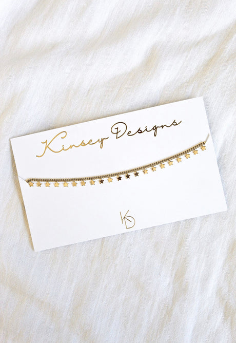 The Starry Choker by Kinsey Designs Archives, a gold-plated 14” necklace with a star charm, is displayed on a white branded card against white fabric for an elegant, stylish look.