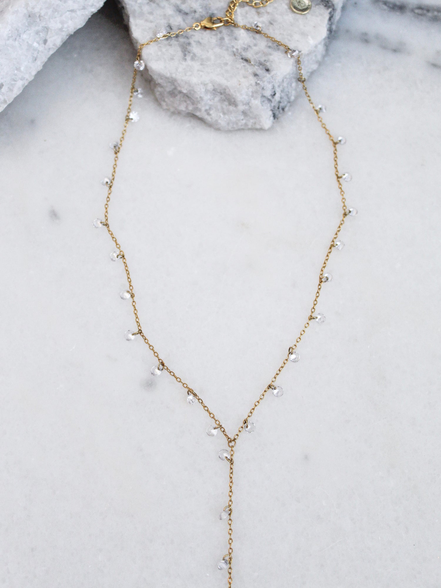 The Clare Lariat by Kinsey Designs Archives is an 18k gold plated necklace with small clear beads, CZ charms, a drop pendant, and an adjustable extender. Shown on marble, it adds elegance to any look.