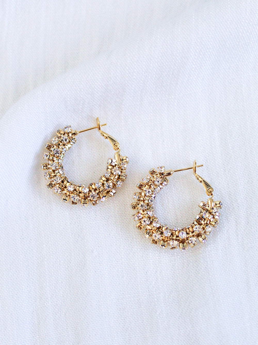 Kinsey Designs' Harkness Hoops, featuring sparkling cubic zirconia, are showcased against a white textured fabric backdrop.