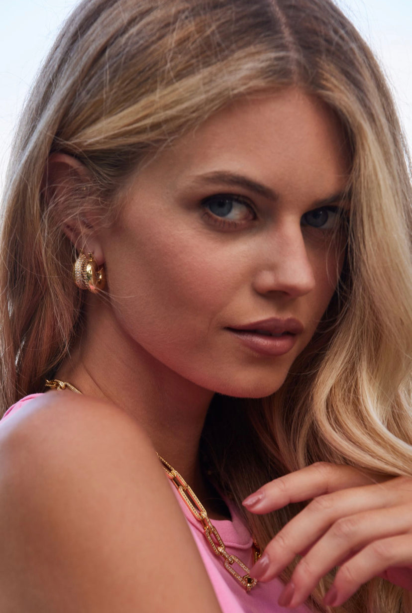 Woman with long blonde hair wears Kinsey Designs Archives' Mikki Pave Hoop gold plated earrings (.75" wide) and a pink top, looking softly at the camera.