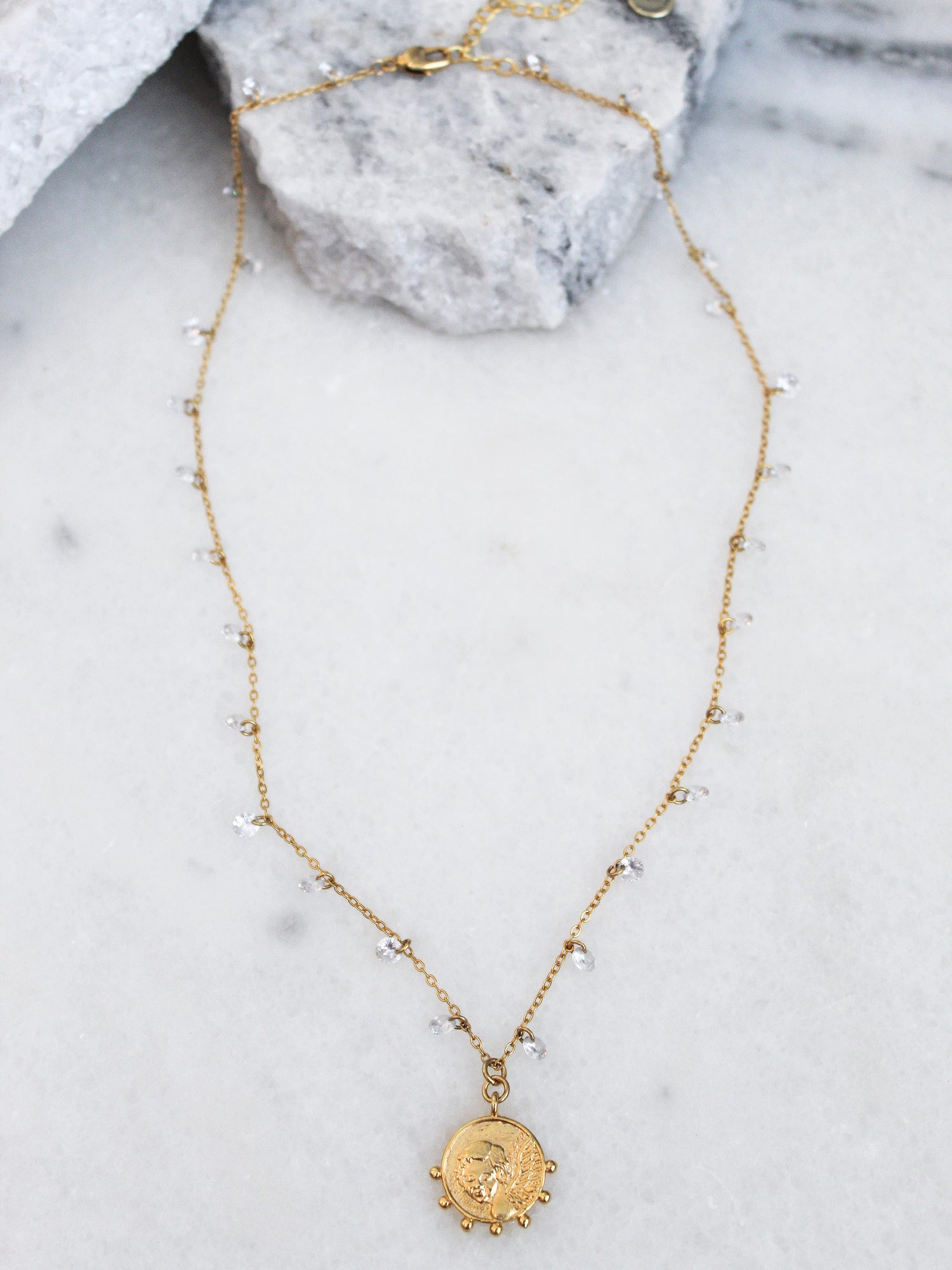The Lover necklace by Kinsey Designs Archives features an 18k gold plated adjustable chain with a round pendant, small beads, and CZ charms. Shown on marble with stone accents, it adds elegance to any look.