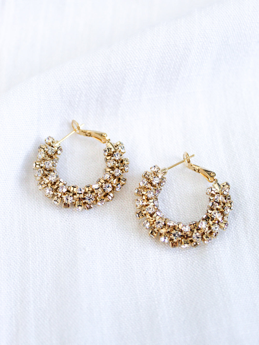 The Harkness Hoops by Kinsey Designs feature gold crystal hoops adorned with clusters of sparkling cubic zirconia, shown against a white fabric background.