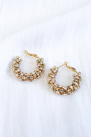 The Harkness Hoops by Kinsey Designs feature gold crystal hoops adorned with clusters of sparkling cubic zirconia, shown against a white fabric background.