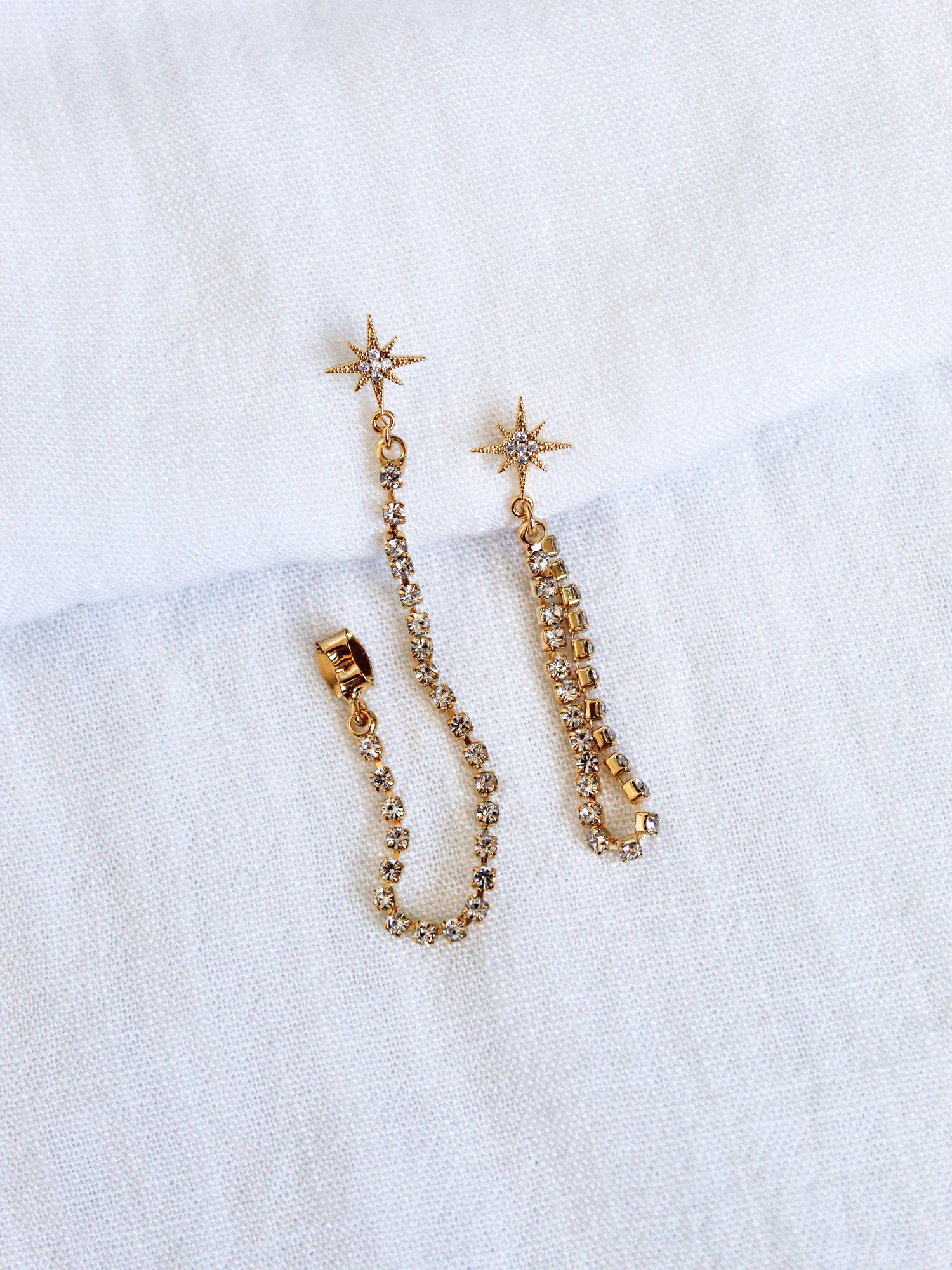 The Reese Drop Earrings by Kinsey Designs Archives are gold plated, star-shaped with crystal chains, shown on a white textured fabric background.