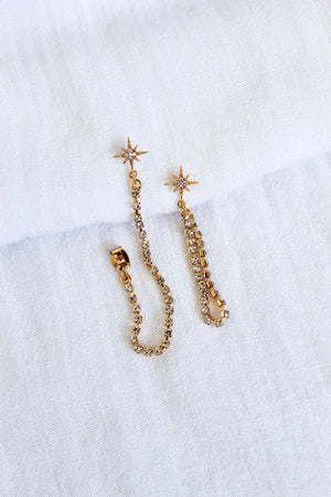 The Reese Drop Earrings by Kinsey Designs Archives are gold plated, star-shaped with crystal chains, shown on a white textured fabric background.