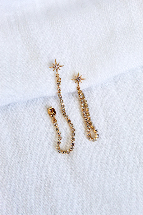 The Reese Drop Earrings by Kinsey Designs Archives are gold plated, star-shaped with crystal chains, shown on a white textured fabric background.