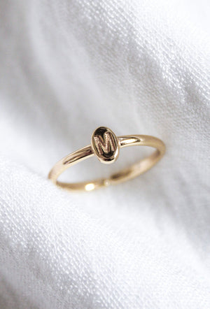 The Kinsey Initial Ring by Kinsey Designs features an oval gold face engraved with the letter "M," displayed on soft white fabric—a timeless, personalized jewelry piece.