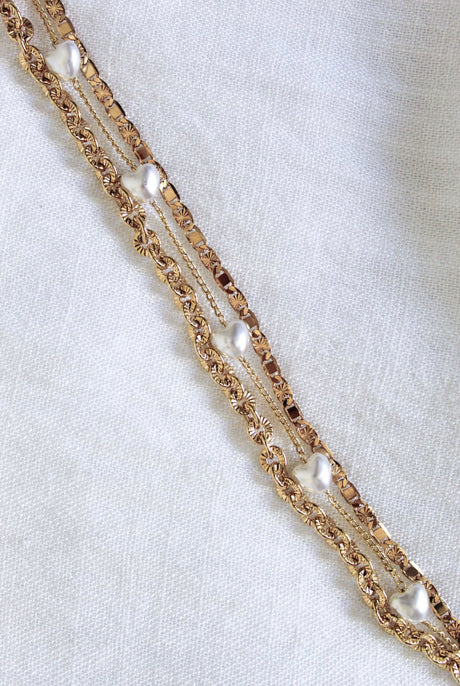 The Beyah Layer Bracelet by Kinsey Designs Archives features gold-plated brass chains with small pearls, displayed diagonally on a white textured fabric background.