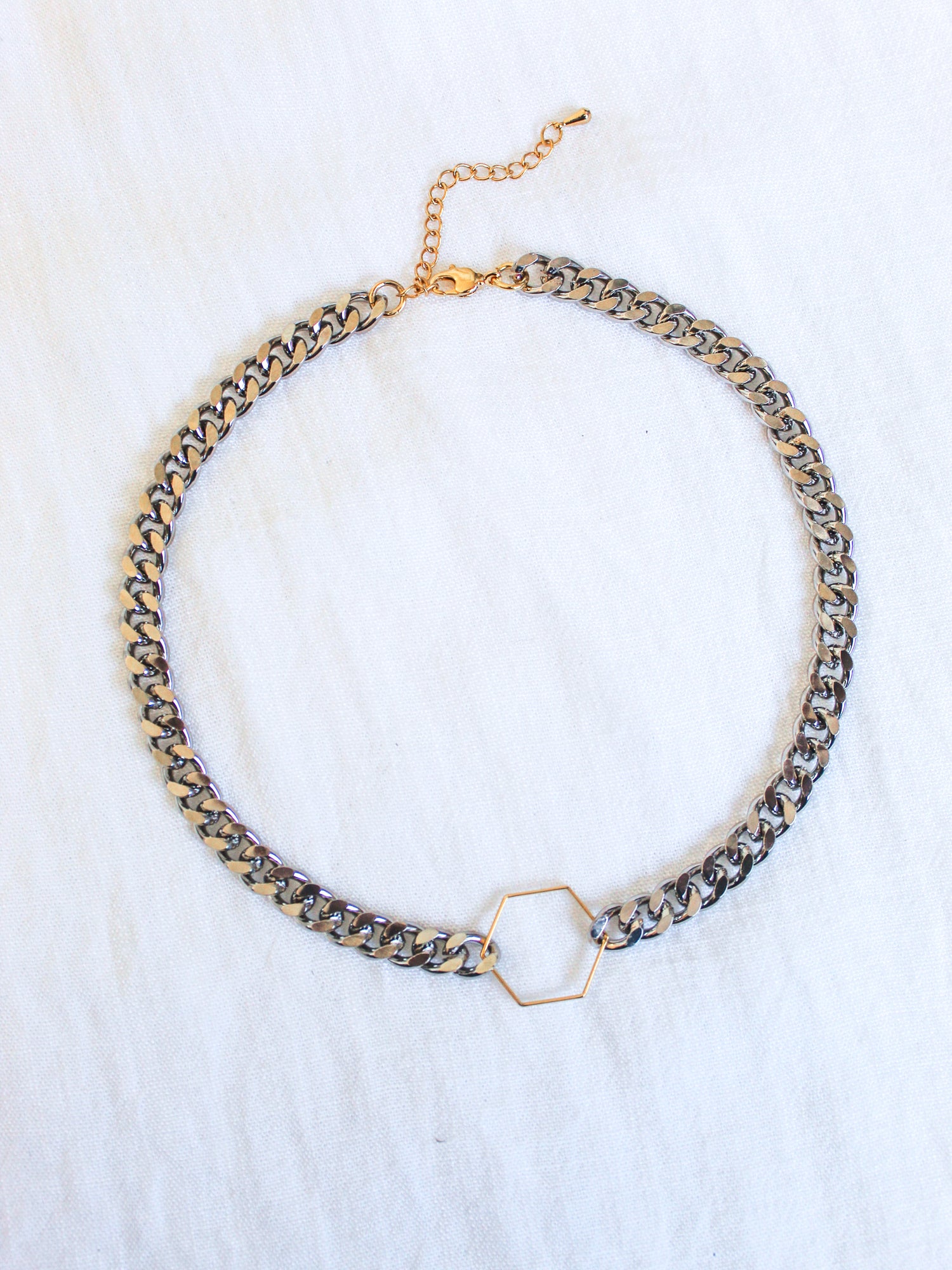 The Fraser Necklace by Kinsey Designs Archives features a 17-inch silver chain with a gold hexagon pendant, crafted from rhodium-plated brass and finished with an adjustable clasp and extender, displayed on white fabric.