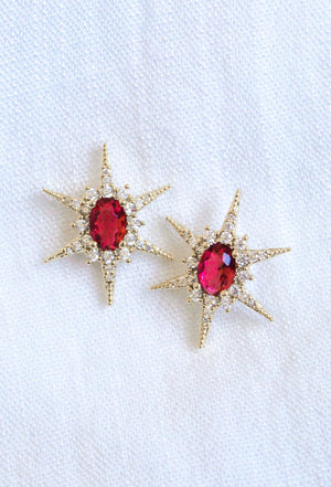 The Faye Stud earrings by Kinsey Designs feature gold-filled starbursts with red gemstone centers and sparkling cubic zirconia accents, shown on white fabric.