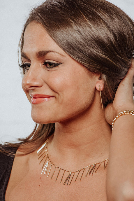 A brown-haired woman smiles, wearing Kinsey Designs Archives’ Walker collection: an 18k gold plated necklace, bracelet, and earrings with surgical steel posts.