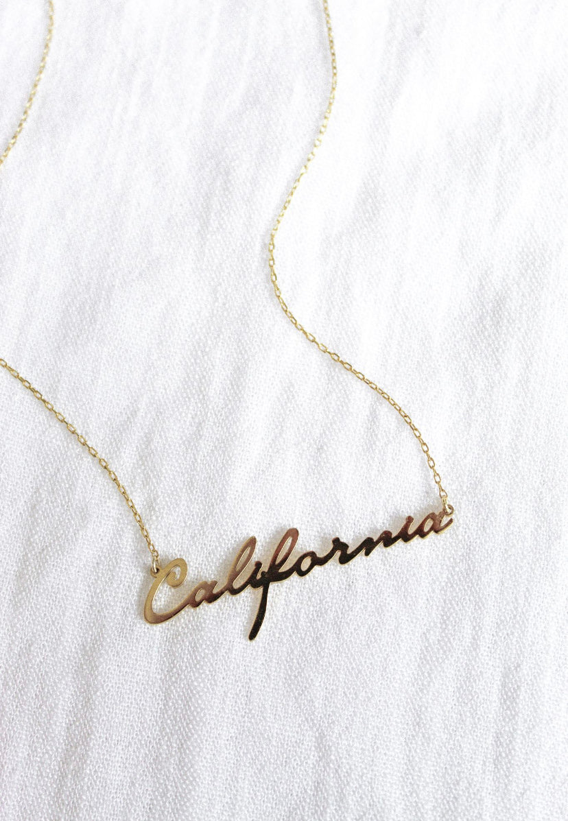 The State of Mind Necklace by Kinsey Designs Archives features a "California" nameplate and is shown on a white textured fabric background.