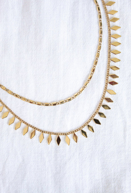 The Fisher Necklace by Kinsey Designs Archives features two layered 14k gold plated geometric pendants, gleaming on a white textured fabric. This modern accessory adds style to any outfit.