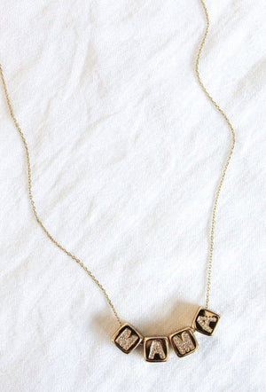 The Kinsey Designs MAMA Block Necklace features gold lettered blocks spelling "MAMA," displayed on a white textured fabric background—a stylish mama necklace and perfect Mother’s Day gift.