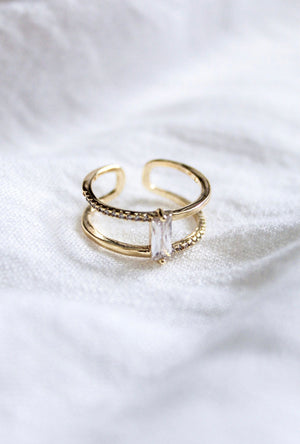 Kinsey Designs' Cora Ring features two bands—one set with small stones and a bold central baguette CZ—shown on white fabric.