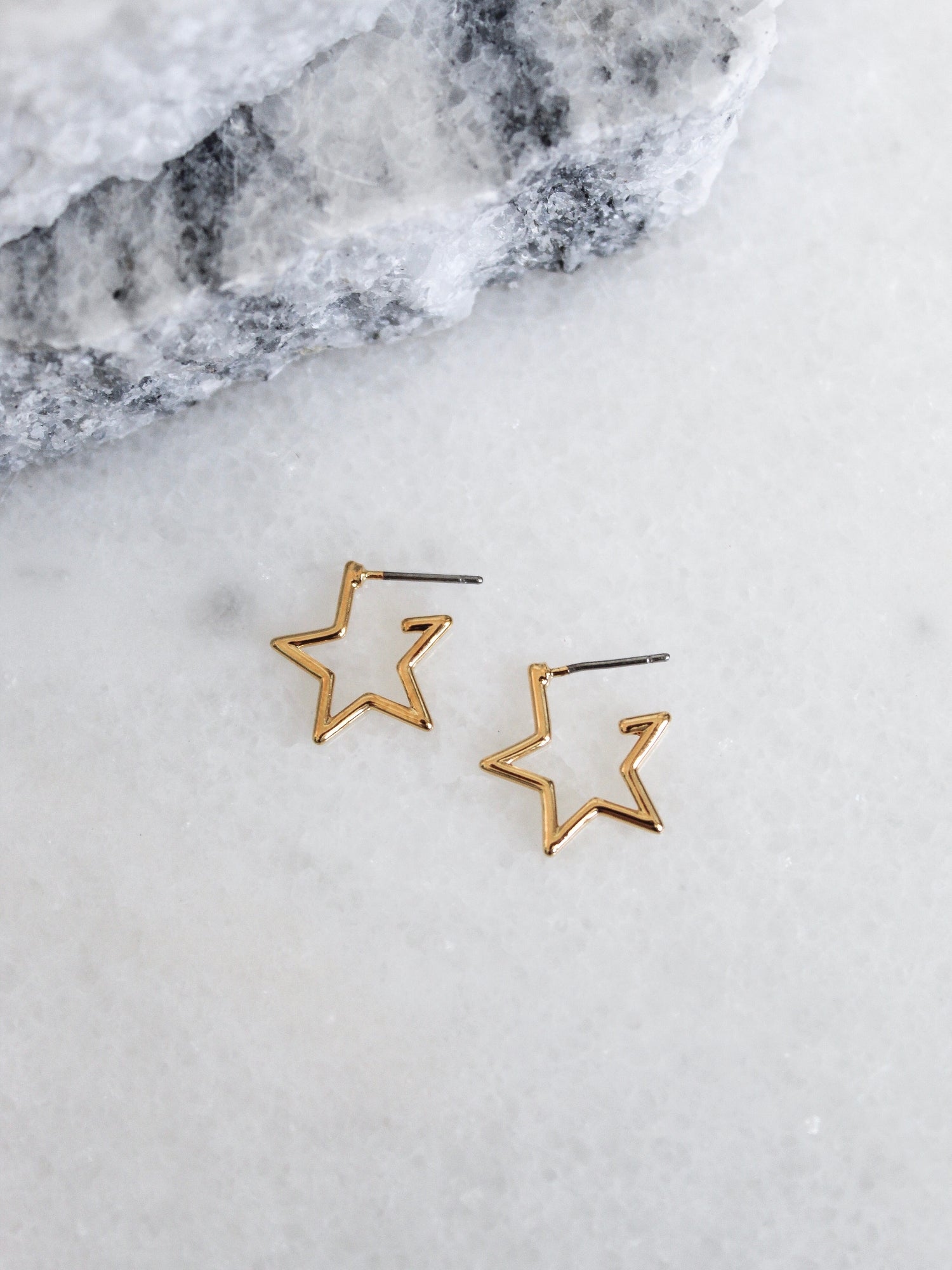 The Starline earrings by Kinsey Designs Archives are gold plated and star-shaped, 0.5” wide, shown on a white marble surface with gray stone in the background.