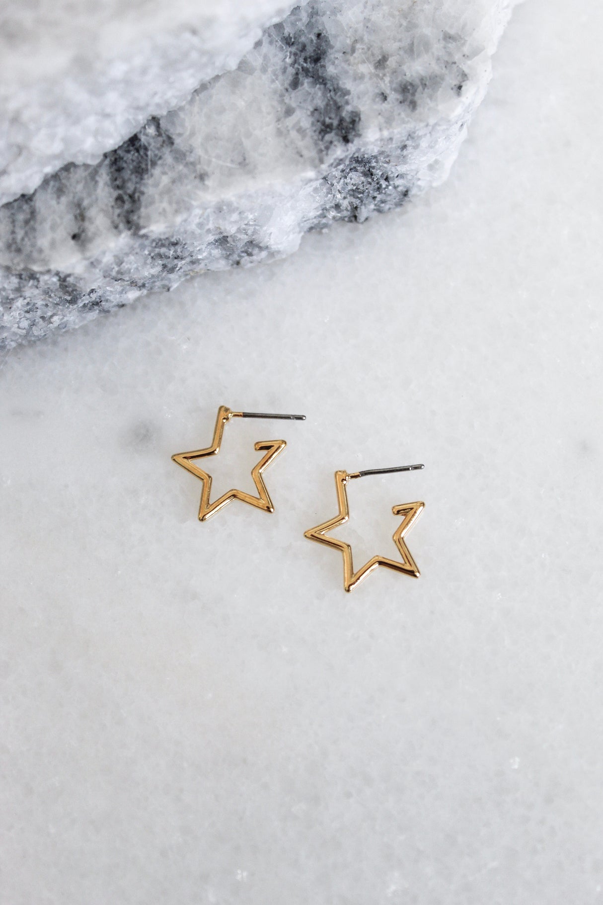 The Starline earrings by Kinsey Designs Archives are gold plated and star-shaped, 0.5” wide, shown on a white marble surface with gray stone in the background.