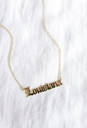 The State of Mind Necklace by Kinsey Designs Archives features a gold-filled hand-stamped "Louisiana" pendant, showcased on a white fabric background.