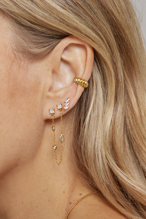 Close-up of a woman's ear adorned with multiple gold-plated and diamond earrings, featuring the Metz Cuff by Kinsey Designs Archives, along with a chain earring and an ear cuff.